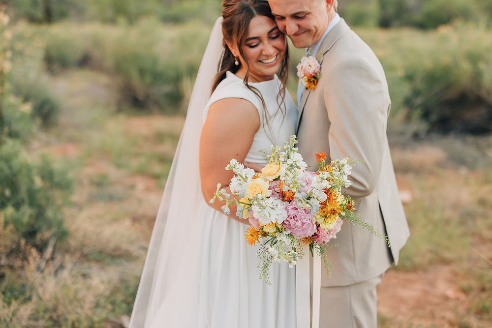Smiling bride holds colorful bouquet while groom embraces her from behind in outdoor desert wedding setting.