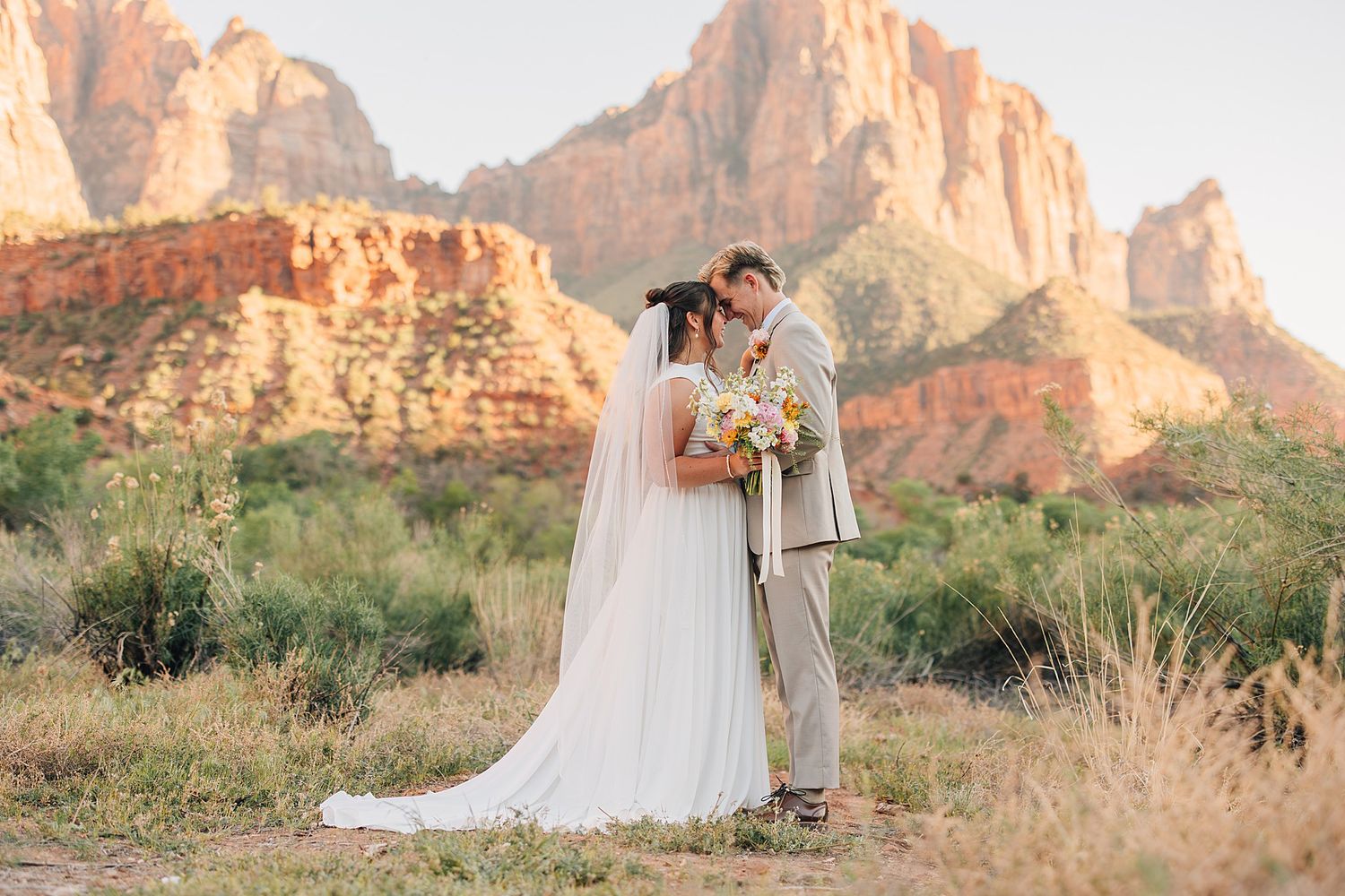 Bride and groom share a kiss at Zion National Park with red rock mountains glowing at sunset.