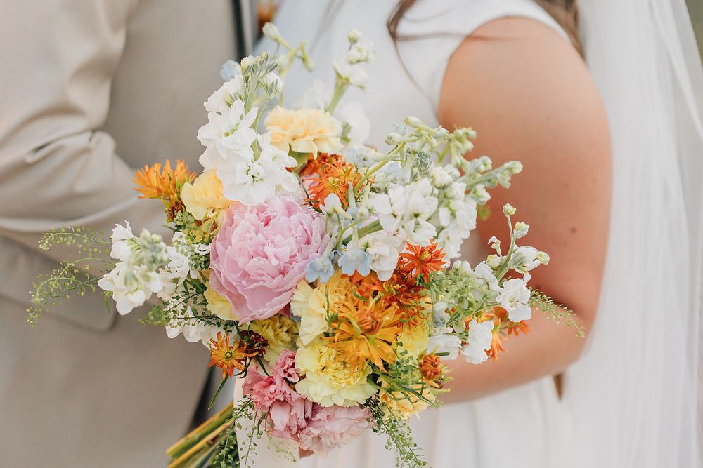 Bride holding a colorful wedding bouquet with pink peonies, orange dahlias, and white flowers.