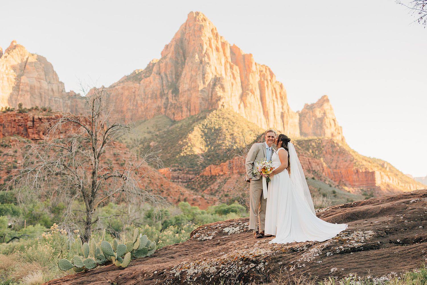Bride and groom kissing at Zion National Park with The Watchman mountain glowing at sunset.