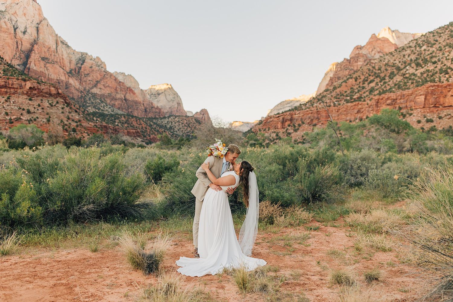 Couple sharing a kiss in wedding attire surrounded by desert shrubs and red rock mountains at Zion National Park.