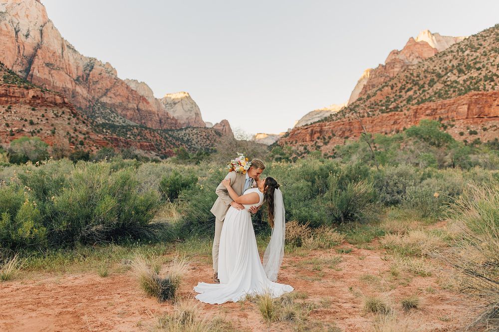 Couple sharing a kiss in wedding attire surrounded by desert shrubs and red rock mountains at Zion National Park.