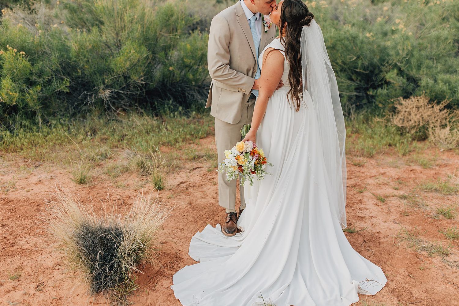 Bride and groom kissing in desert landscape, bride holding colorful bouquet in flowing white gown with veil.