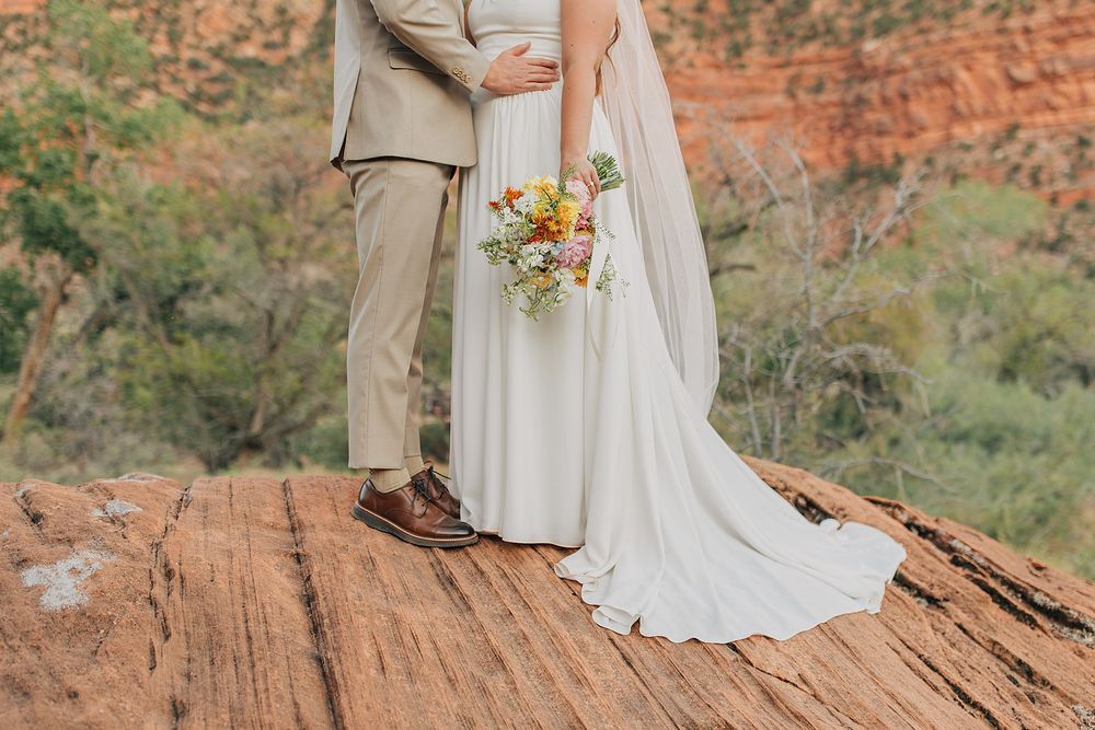 Bride and groom standing on red rock cliff, bride holding wildflower bouquet in white gown with train.