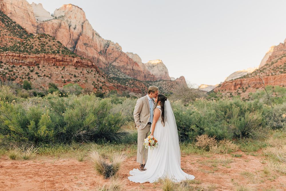 Bride and groom kissing at Zion National Park with red rock canyon mountains and desert landscape backdrop.