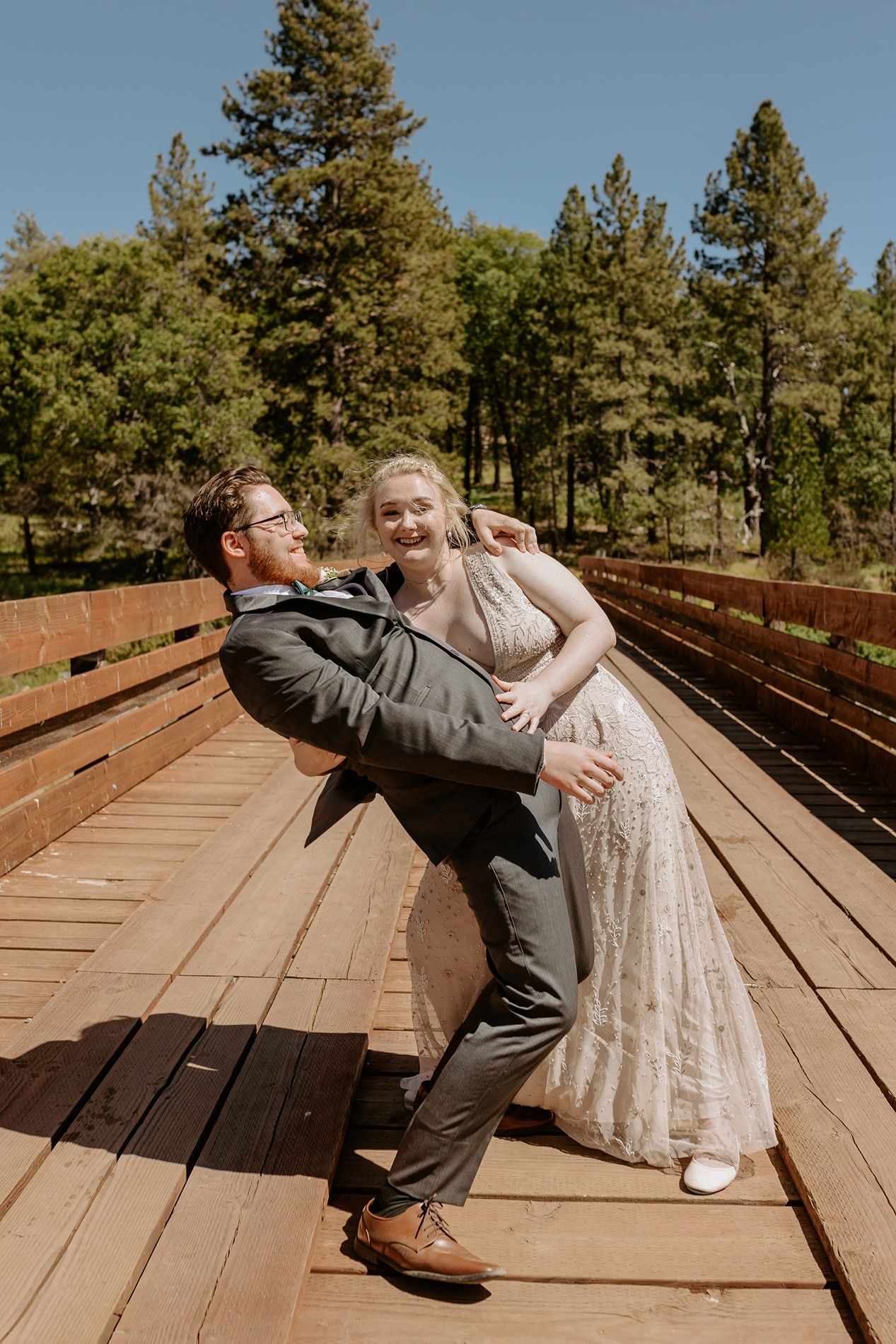 Joyful couple sharing a playful moment on a wooden bridge surrounded by tall pine trees on their wedding day at Lake Cuyamaca, Julian.
