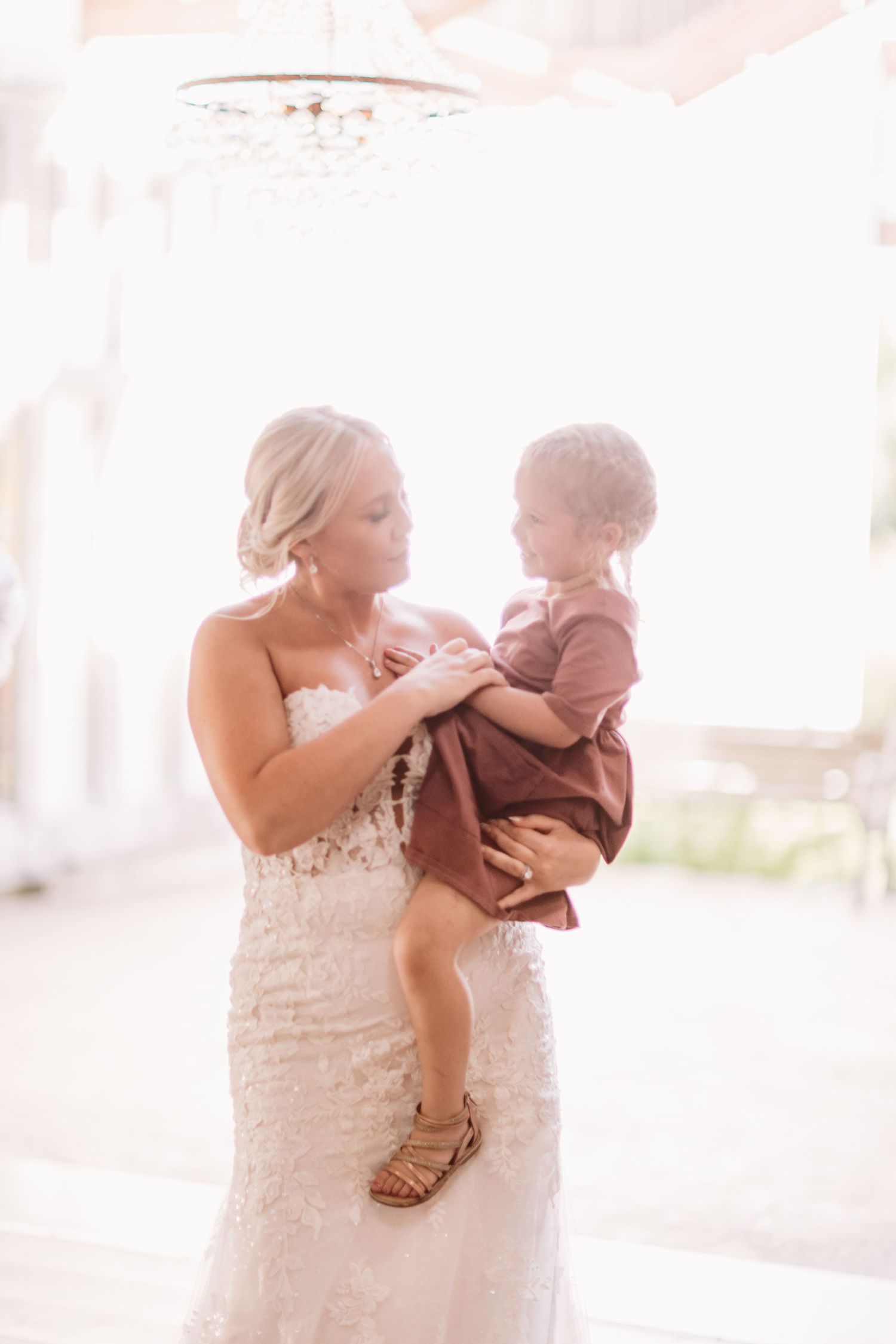Bride in lace wedding gown holds young child in rustic brown outfit, sharing a tender moment in soft natural light.