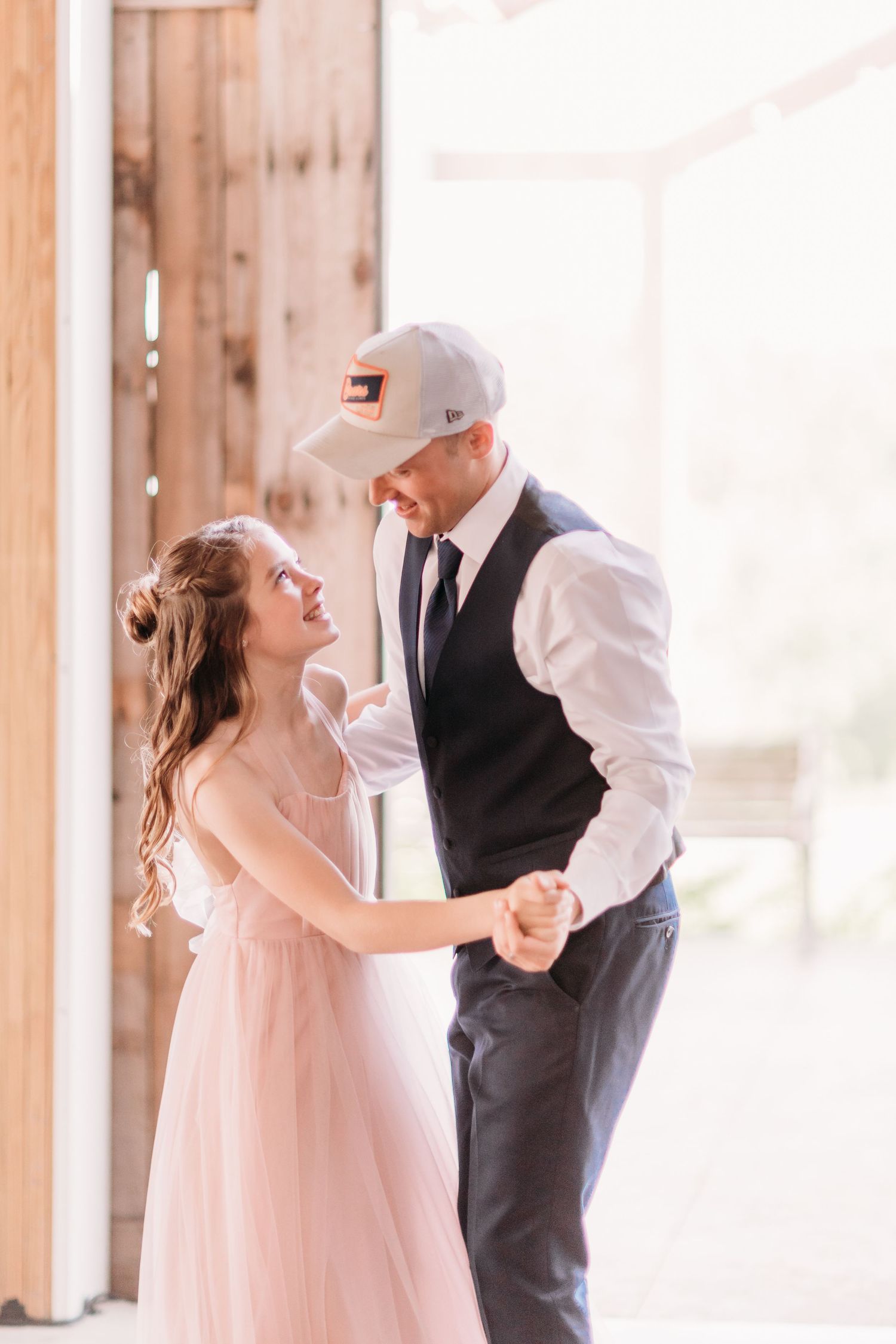 Couple dancing together in a rustic barn venue, woman in pink dress smiling up at man in vest and baseball cap.