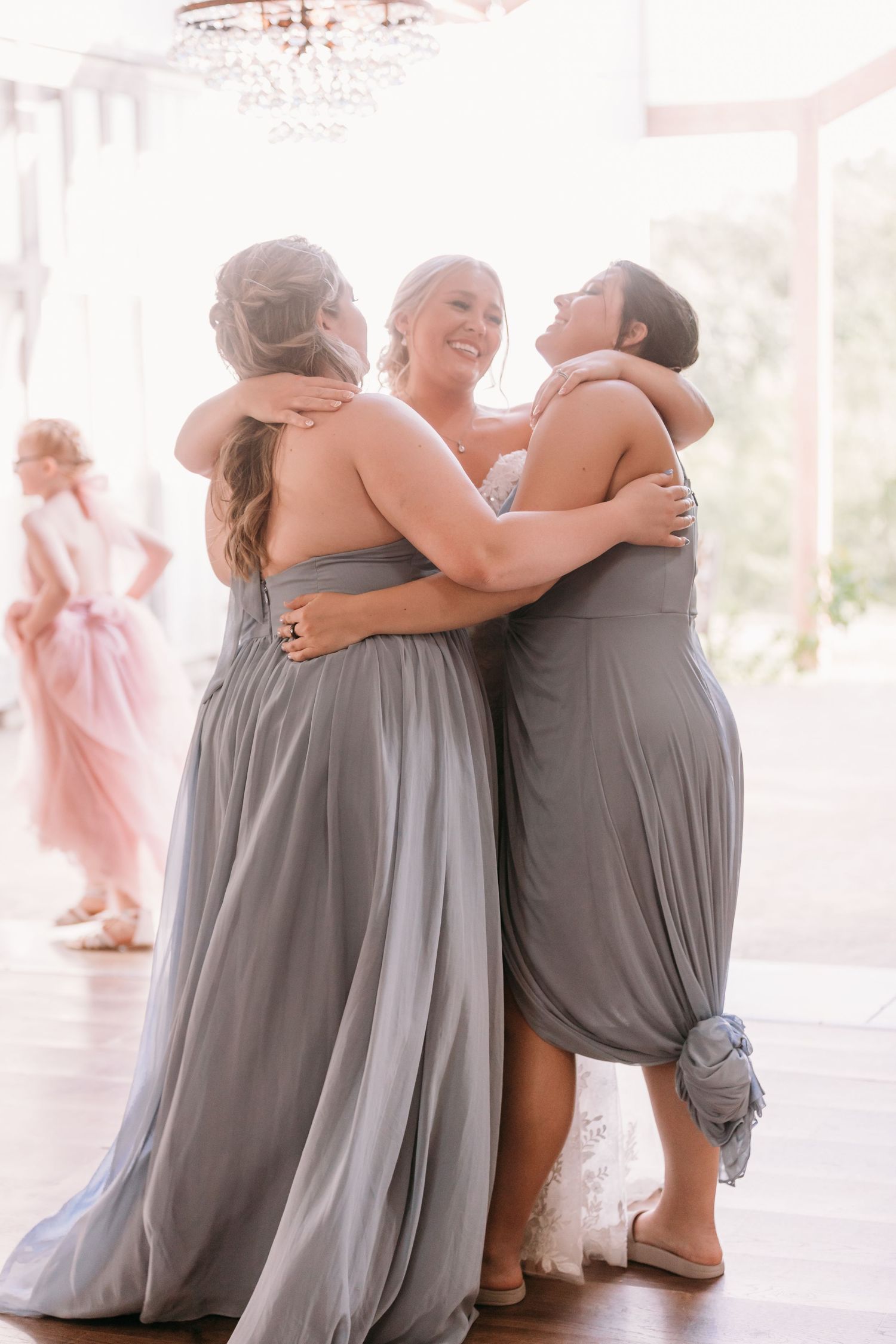 Bride in white gown shares joyful group hug with two bridesmaids in gray dresses at elegant wedding venue.