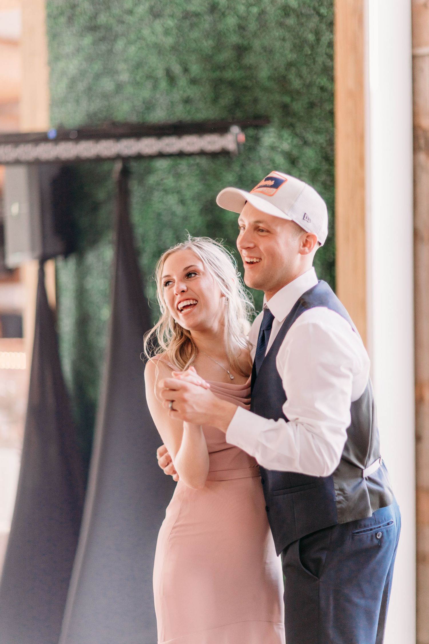 A laughing couple dances together at a wedding reception, woman in pink dress, man in vest and white cap.