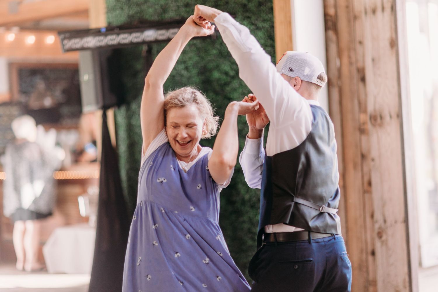 Joyful woman in blue dress being twirled by partner in vest during outdoor wedding reception dance.