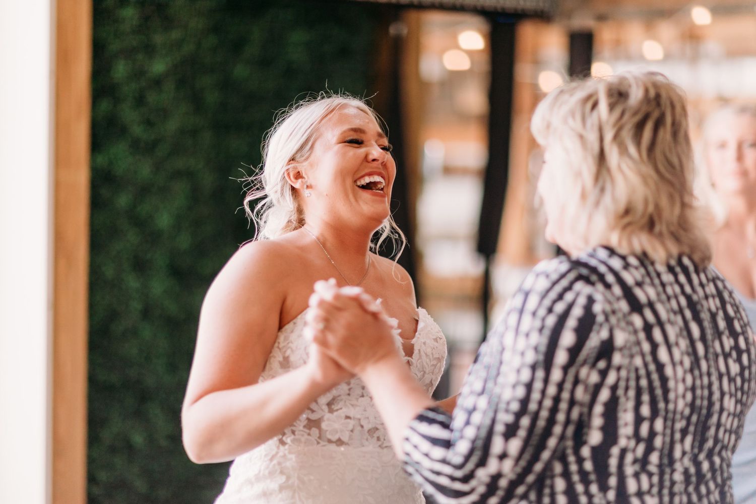 Bride in lace wedding dress laughing and holding hands with guest during joyful wedding reception dance.