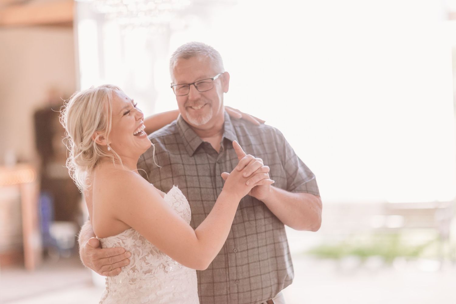 Bride in white dress dancing and laughing with older man in plaid shirt at wedding reception.