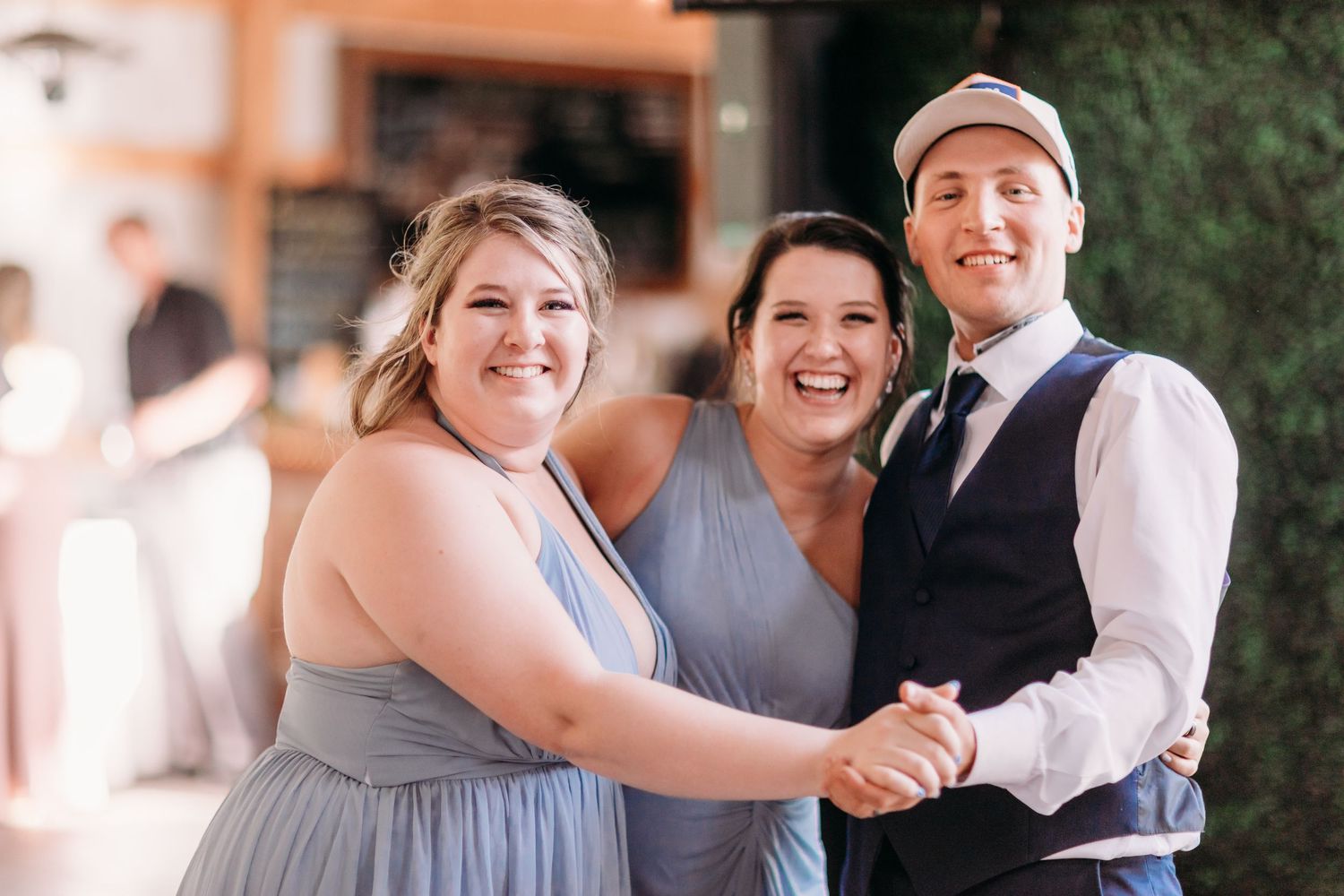 Two bridesmaids in blue dresses smiling with groom in vest at wedding reception.