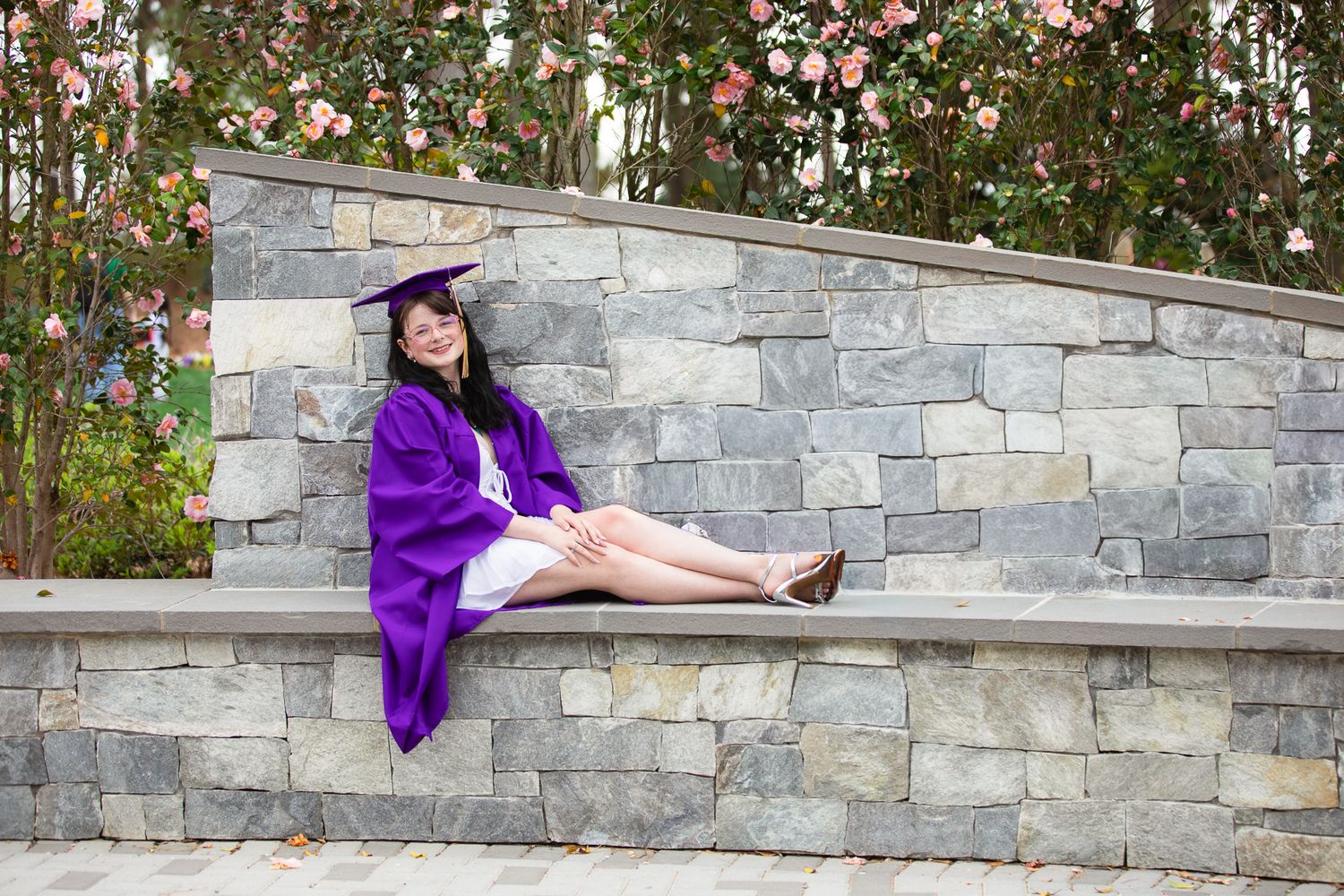 A smiling graduate in purple cap and gown sits on a stone wall surrounded by blooming flowers outdoors at WRAL Azalea Garden