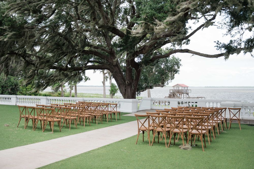 Outdoor wedding ceremony setup with wooden chairs on green lawn under a massive oak tree overlooking the waterfront.