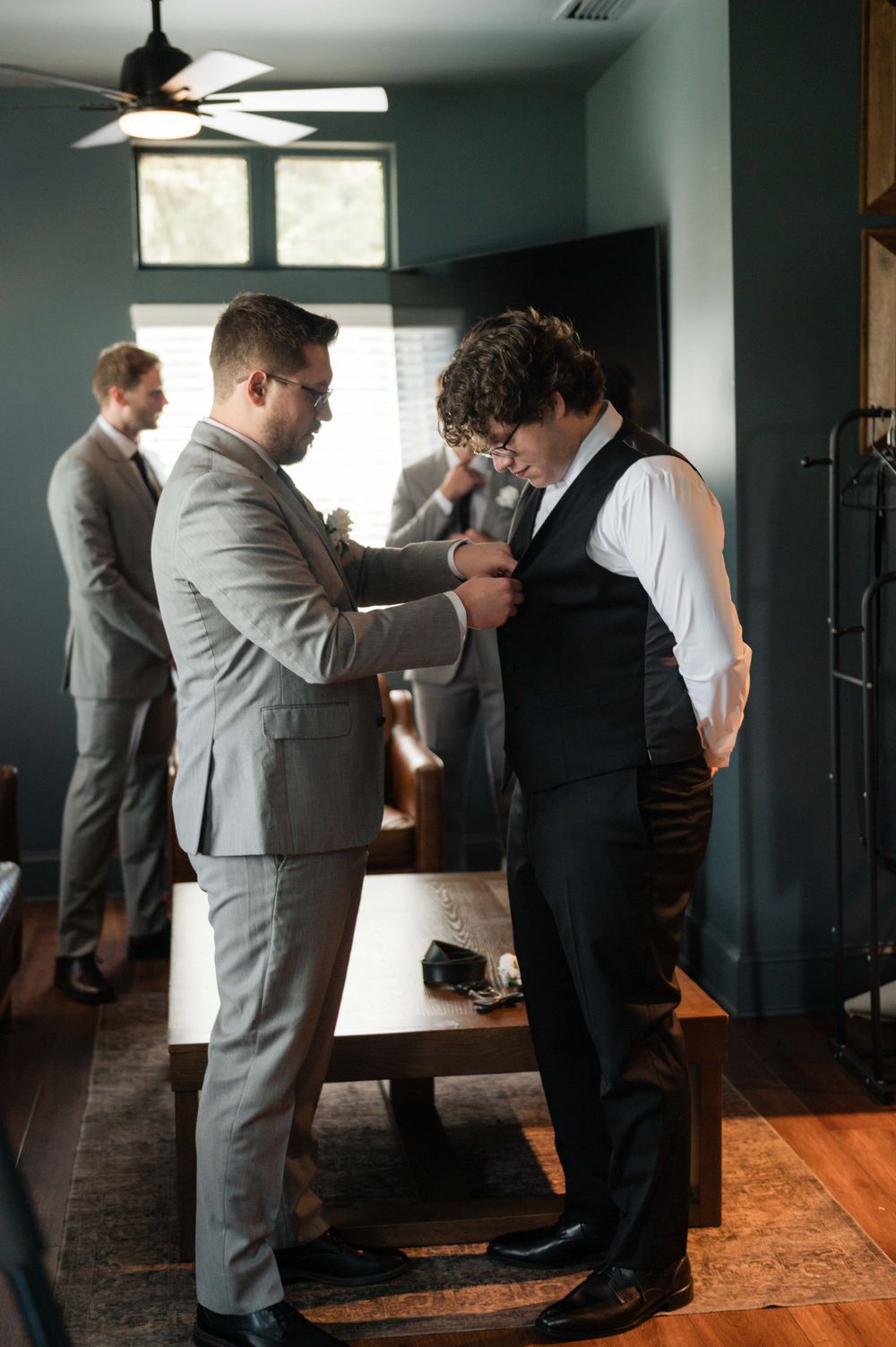 Groomsmen helping each other get dressed in gray suits before a wedding ceremony indoors.