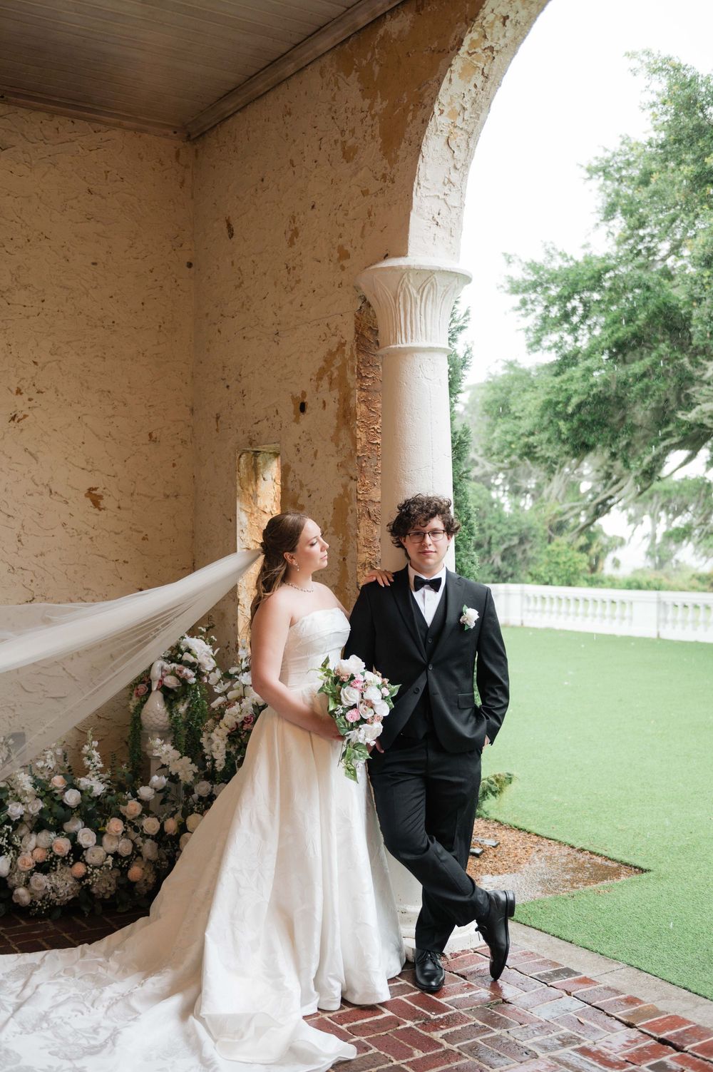 Bride and groom pose under stone arch, her veil flowing in the wind, holding a pink bouquet.