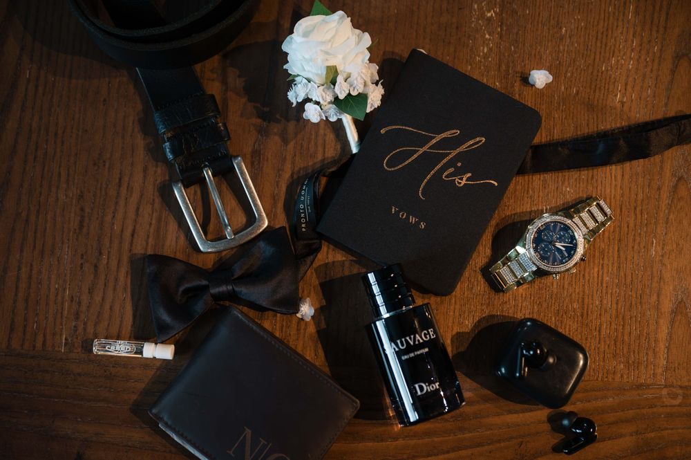 Groom's wedding flat lay featuring a black belt, bow tie, Dior Sauvage cologne, watch, wallet, and floral boutonniere on wood.