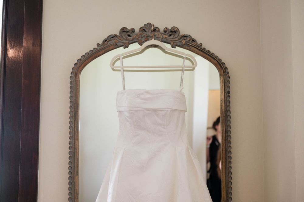 White strapless wedding dress hanging on a vintage ornate mirror with elegant gold frame in a bridal suite.