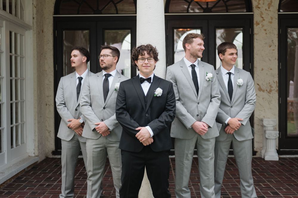 Groom in black tuxedo stands with groomsmen in gray suits and black ties at an elegant wedding venue.