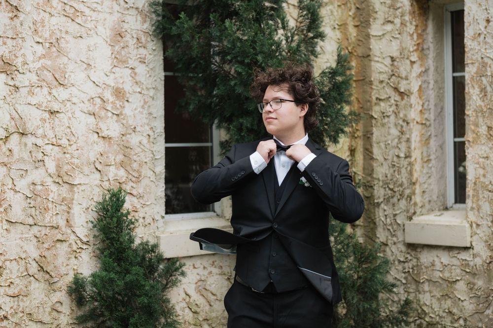 Young person in black suit adjusting tie outdoors near stone building with evergreen shrubs.