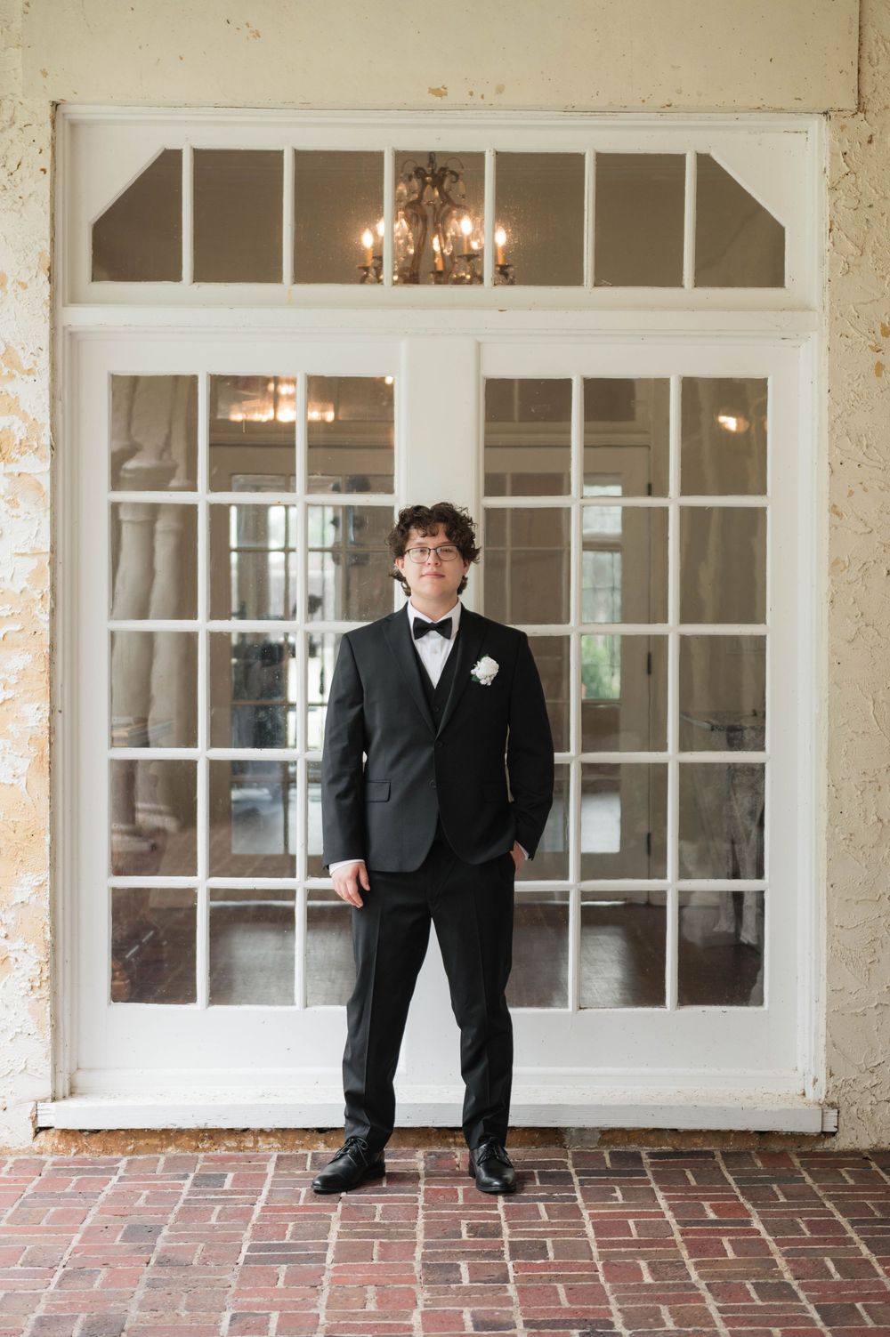 Groom in black tuxedo with bow tie and boutonniere standing before elegant French doors with chandelier.