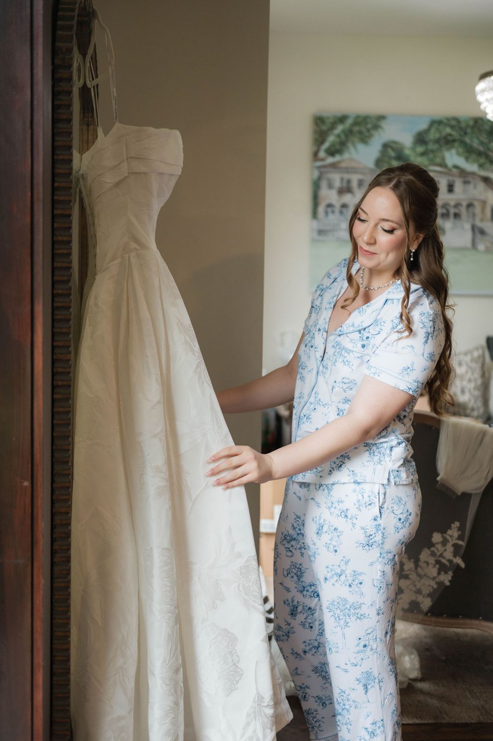 Bride in blue floral pajamas admiring her elegant white wedding gown hanging in a sunlit room.
