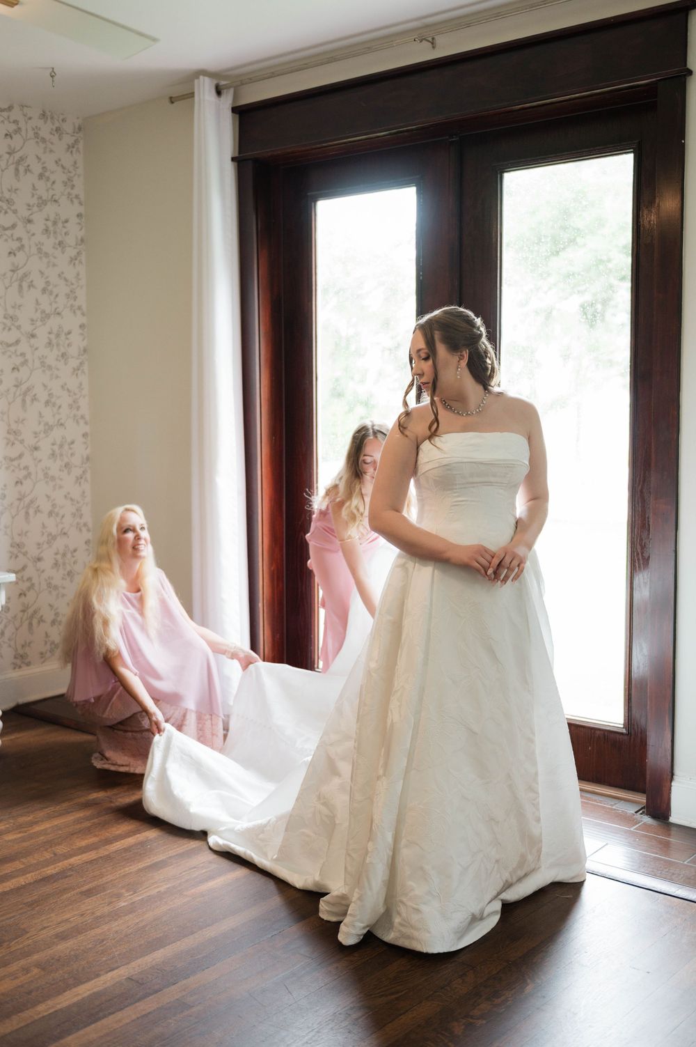 Bridesmaids in pink dresses help adjust the train of the bride's strapless white ball gown.