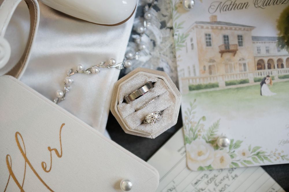 Wedding rings in velvet box with diamond bracelet, bridal shoes, and illustrated wedding invitation on elegant white background.
