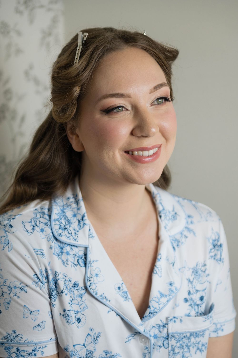 Smiling bride in blue floral pajamas with rhinestone hair clip and elegant makeup during wedding morning preparations.