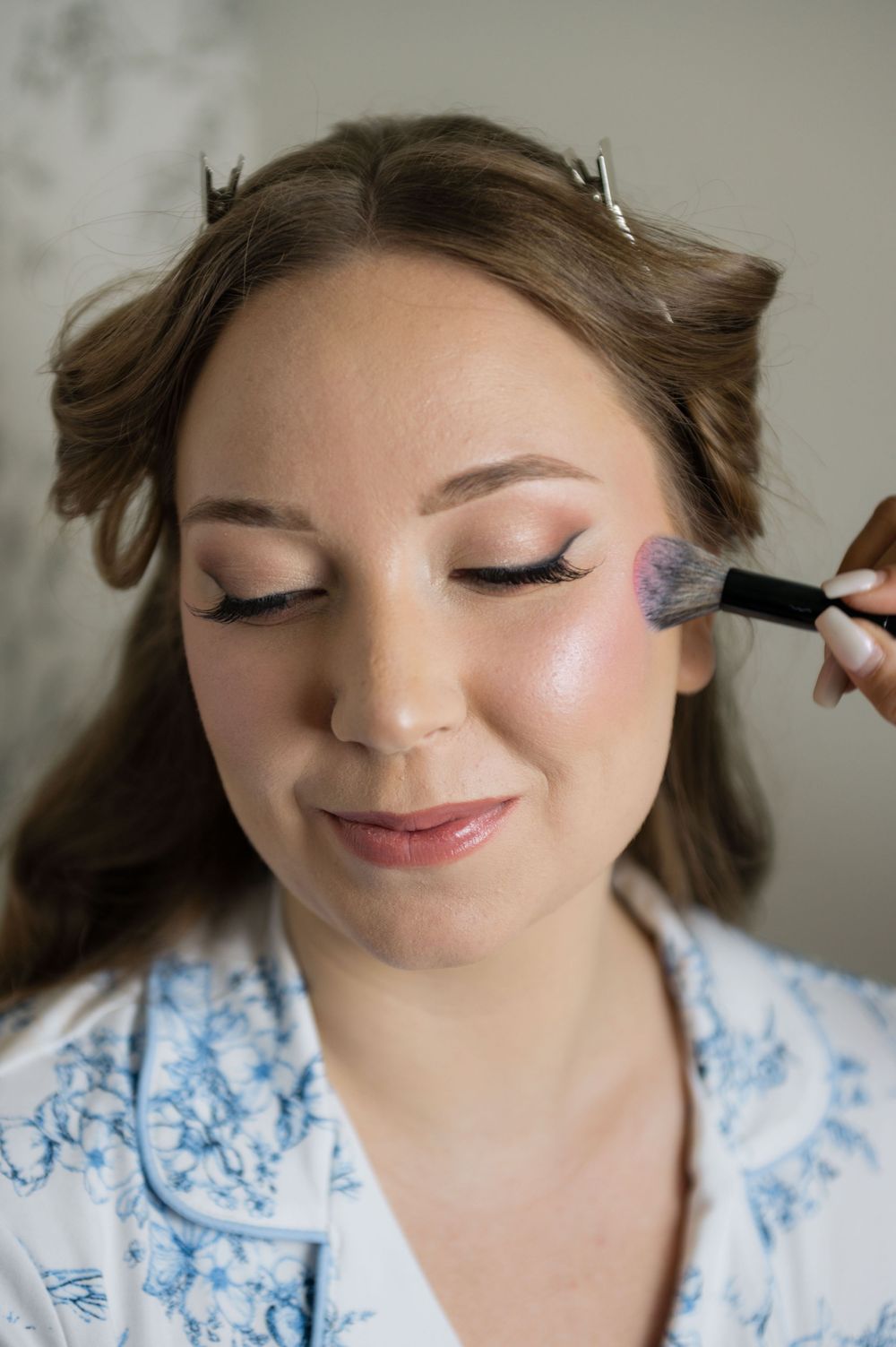 A makeup artist applies blush to a young woman with curled hair wearing a floral robe.
