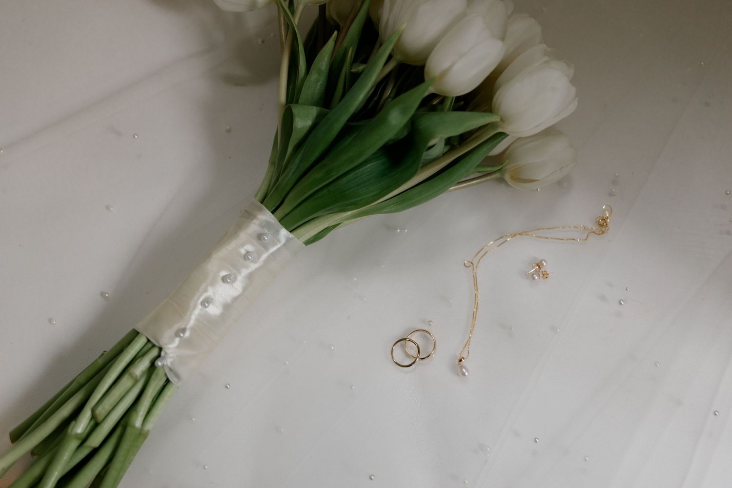White tulip bridal bouquet with satin ribbon beside gold wedding rings and delicate necklace on white surface.