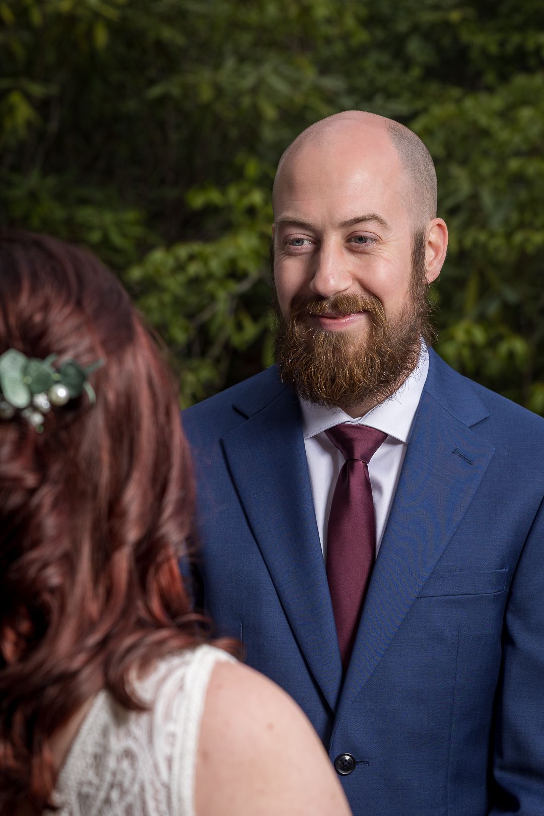 Groom in blue suit and burgundy tie smiling at bride during outdoor wedding ceremony.
