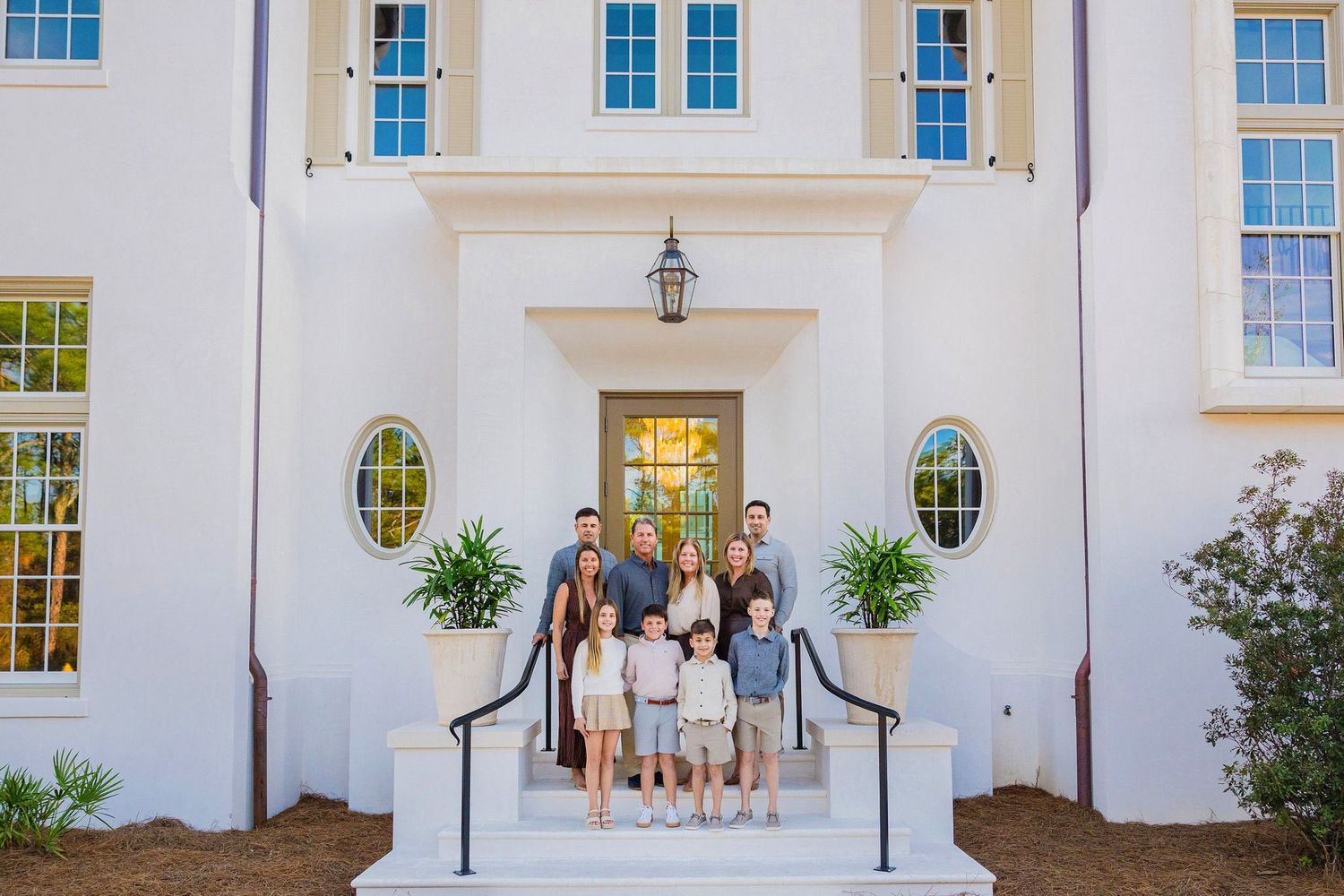 Large family posing on front steps of elegant white two-story home with blue shutters and manicured entrance.
