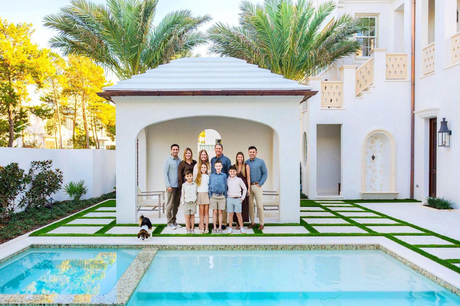 Large family posing together by a luxury pool in front of an elegant white villa with palm trees.