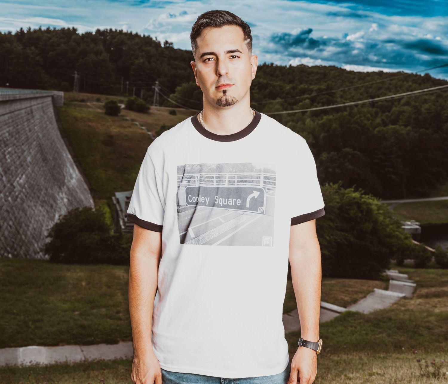 Young man in white graphic ringer t-shirt posing outdoors near a dam with green hills and cloudy sky.