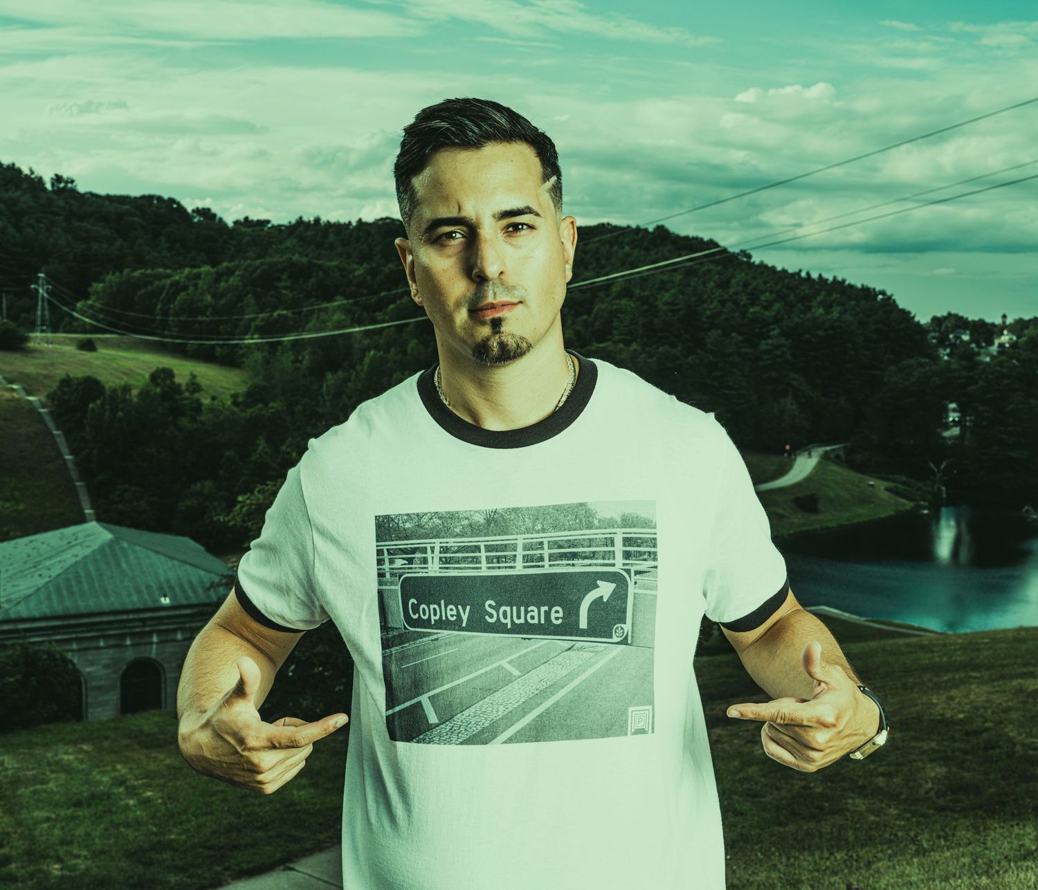 Man in Copley Square t-shirt posing outdoors with green hills and water reservoir in background.