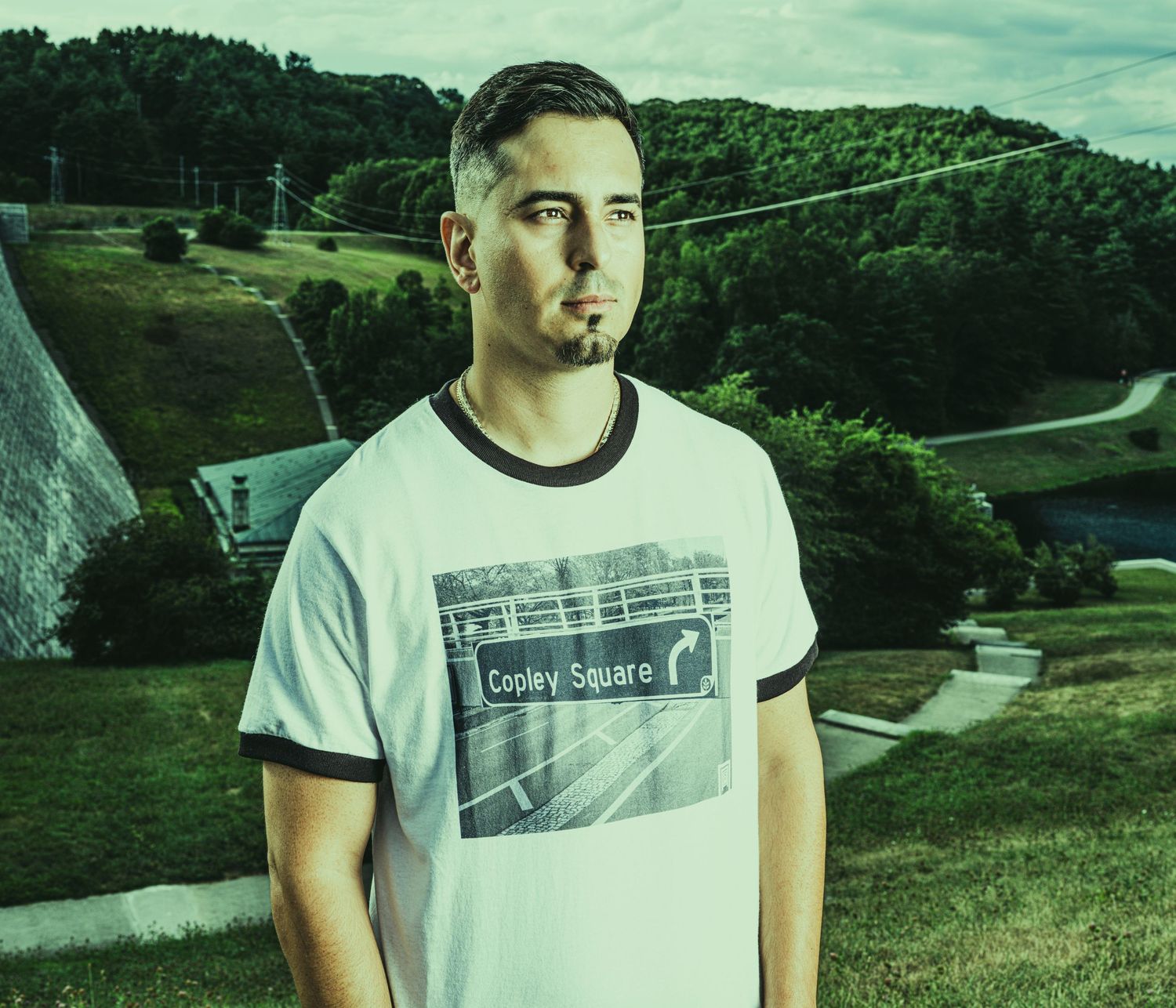 Young man in a Copley Square t-shirt standing outdoors on a green hillside with trees and structures in background.