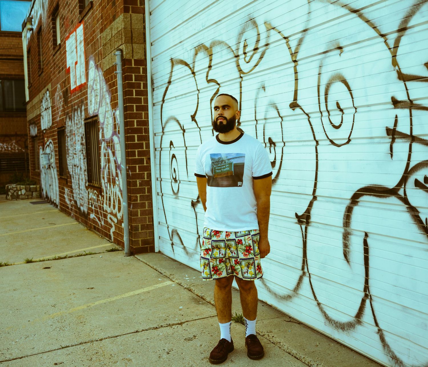 Man in graphic tee and floral shorts stands before graffiti-covered white wall in urban alleyway.