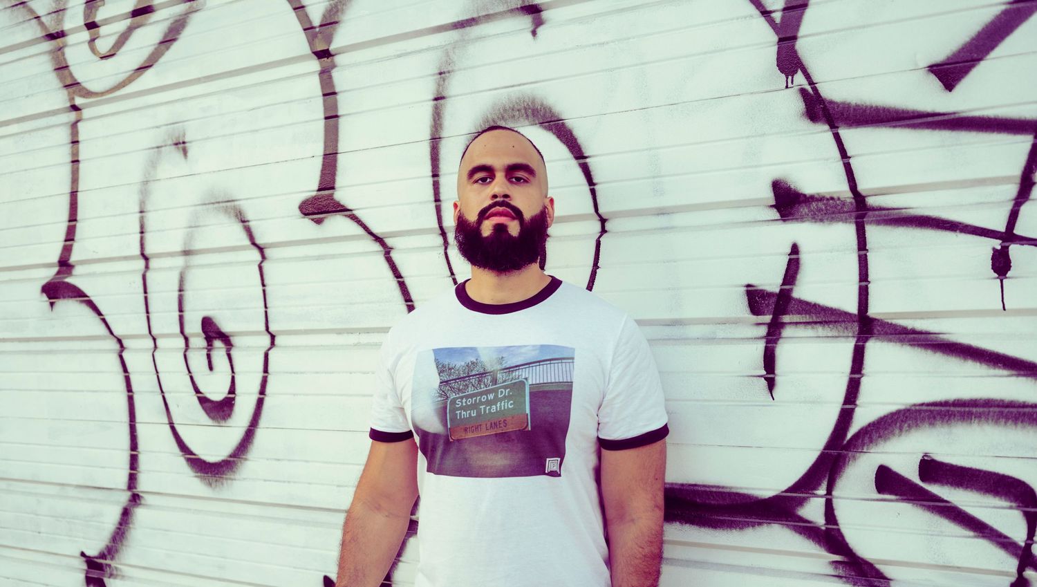 Bearded man in white graphic tee posing confidently against a graffiti-covered white brick wall outdoors.