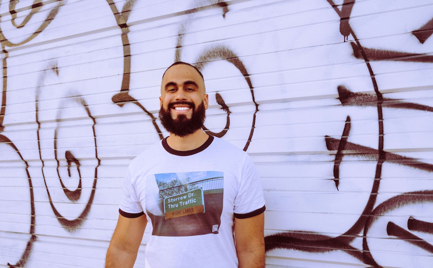 Smiling bald man with beard wearing graphic white t-shirt standing in front of graffiti wall.
