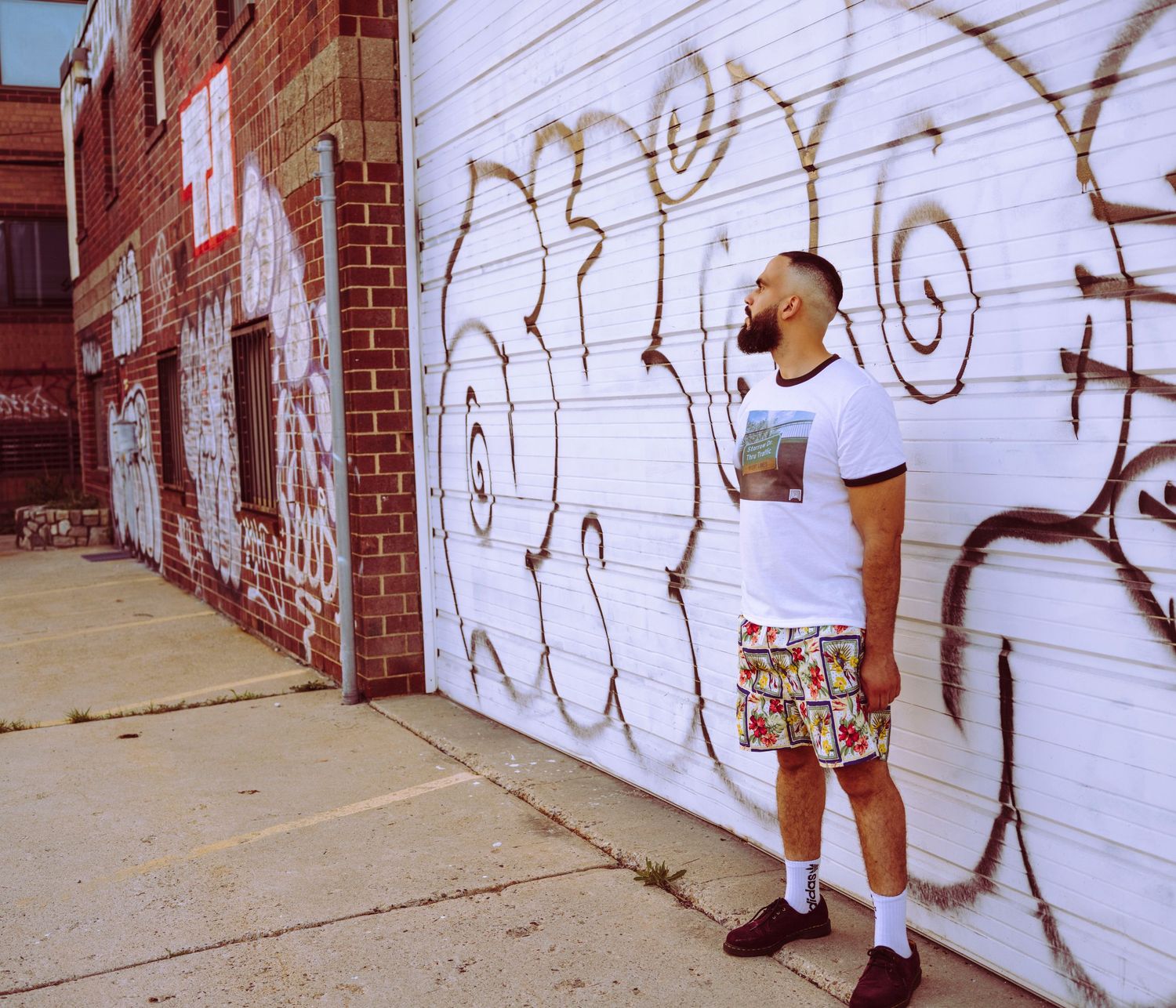 Man in white tee and colorful shorts stands before graffiti-covered white wall on urban brick building sidewalk.
