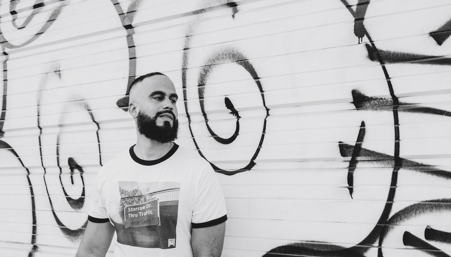 Black and white photo of a bearded man in a graphic tee posing against a graffiti-covered wall.