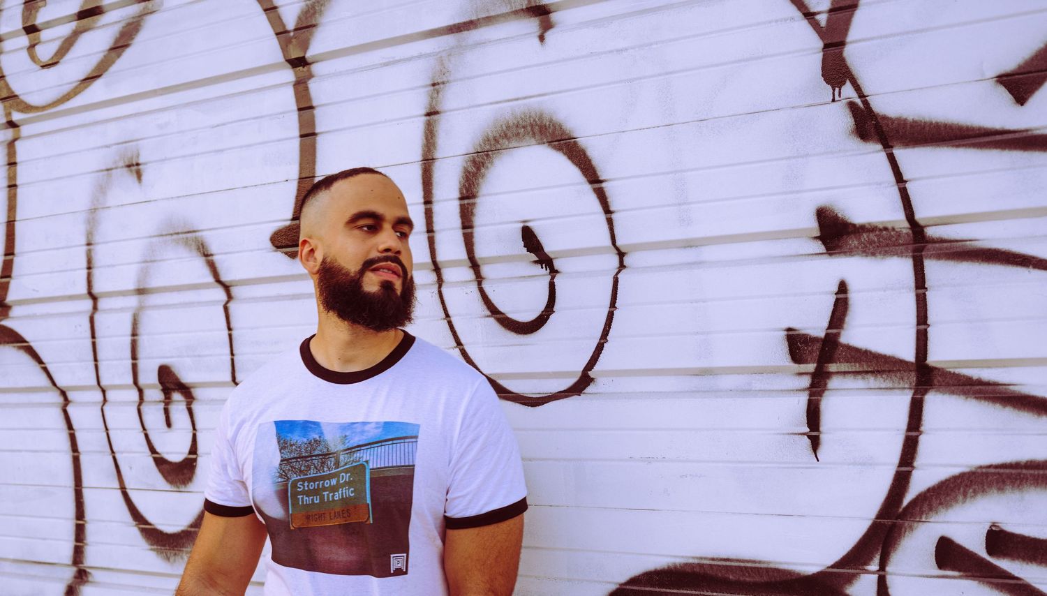 Bearded man in graphic tee posing against white wall with graffiti art outdoors.