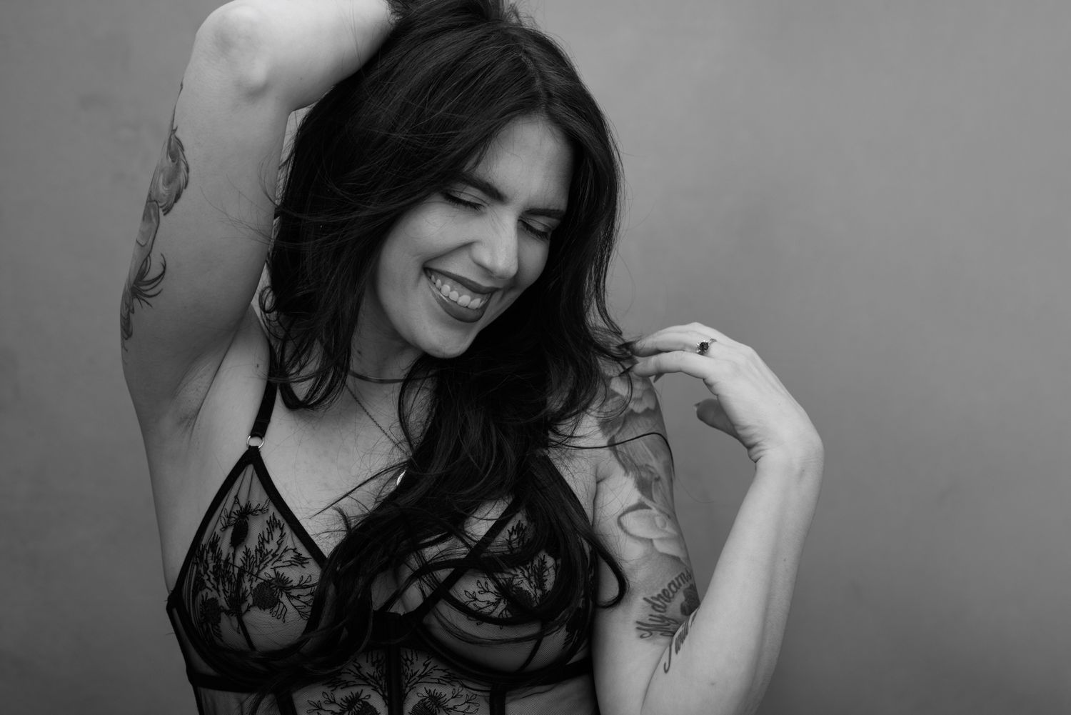 Black and white photo of a smiling tattooed woman with long dark hair posing in lace lingerie.