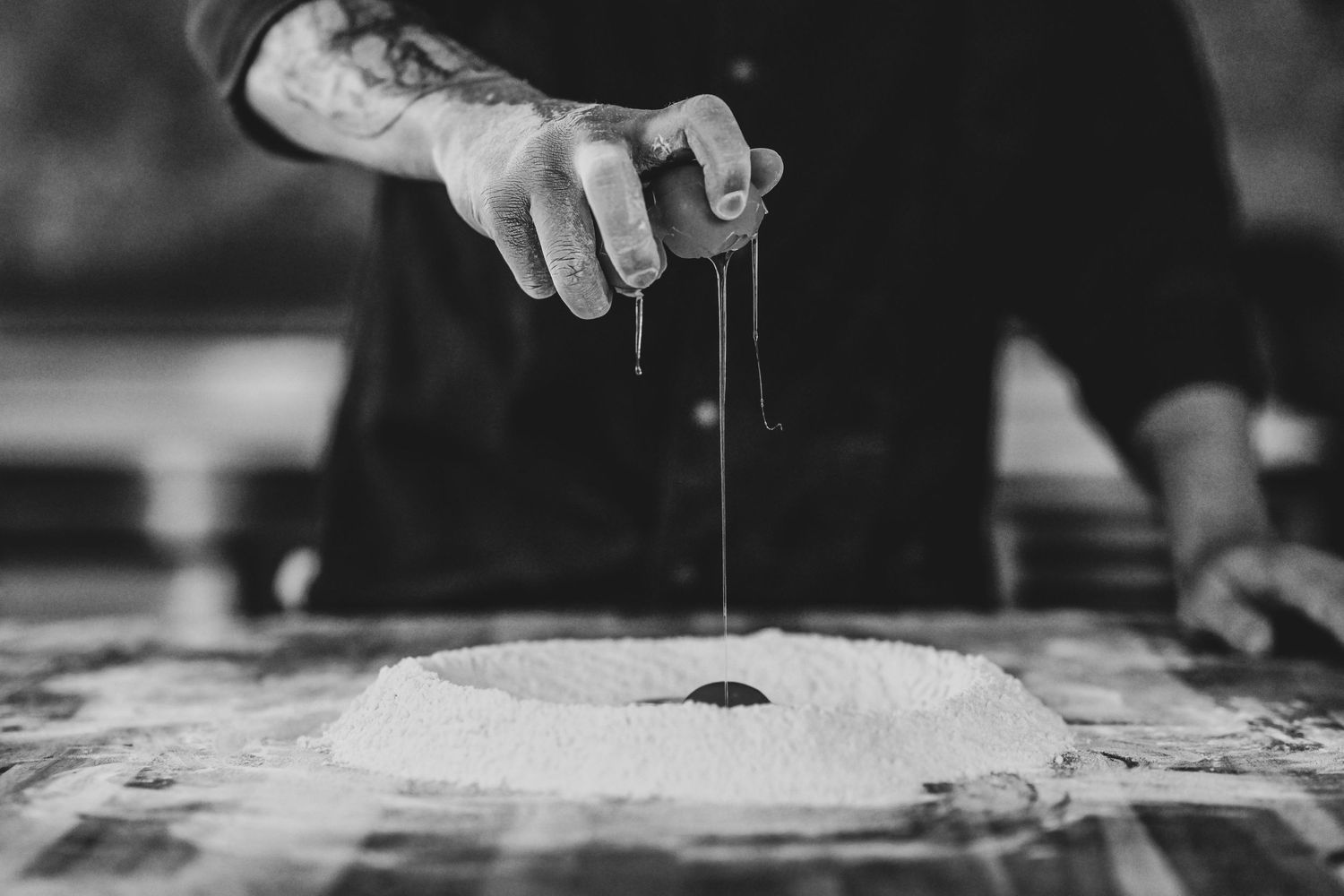 Black and white photo of tattooed hands sprinkling ingredients onto flour mound on a table.