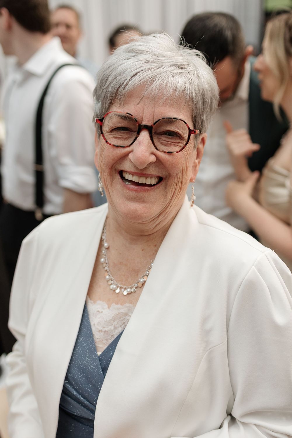 Smiling elderly woman with short gray hair, glasses, and pearl necklace wearing a white blazer at a wedding reception.