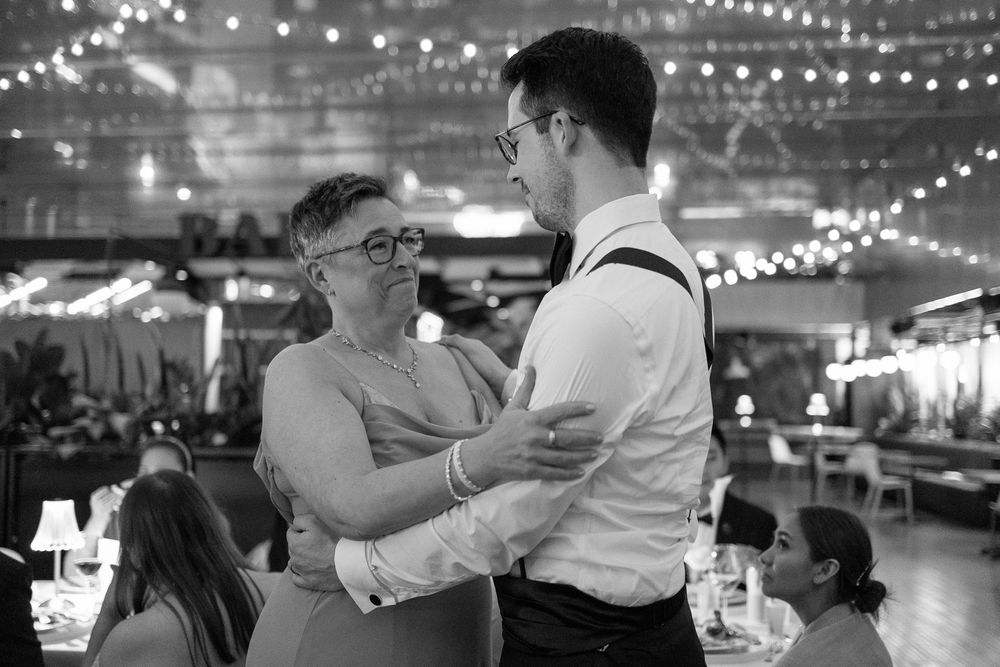 Black and white photo of a mother and son sharing a dance at a wedding reception with string lights above.