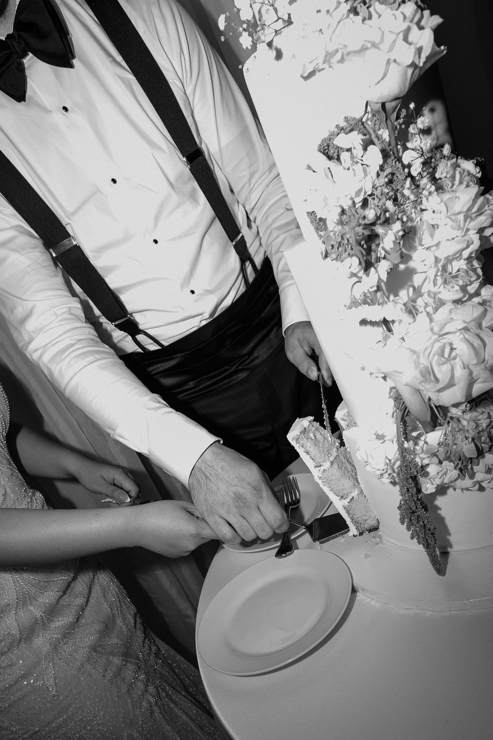 Black and white photo of bride and groom cutting wedding cake, groom wearing suspenders and bow tie.